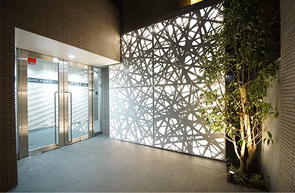 写真：ビル内のオフィス入口。入口正面の壁際には細い木が植樹してあり、下から照明が当たっている。正面入って左側の壁は光が透けていて縦横斜めに色々な幅の線の影が写っている