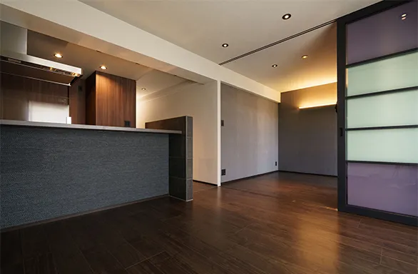 写真：マンション室内。濃い茶色のフローリング床、白い壁、カウンターキッチンは濃い灰色で落ち着いた感じになっている