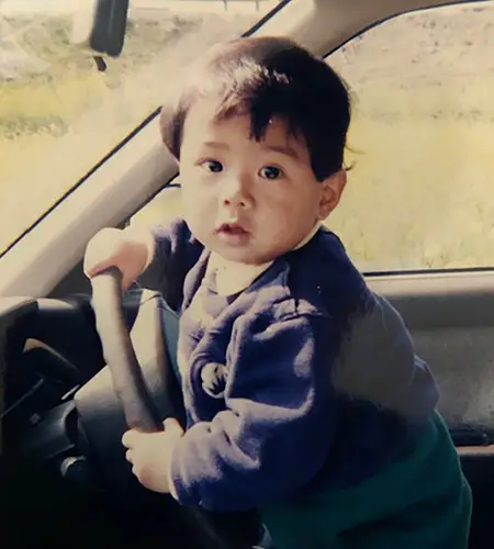 写真：2〜3歳ぐらいの頃で車のハンドルを握っている